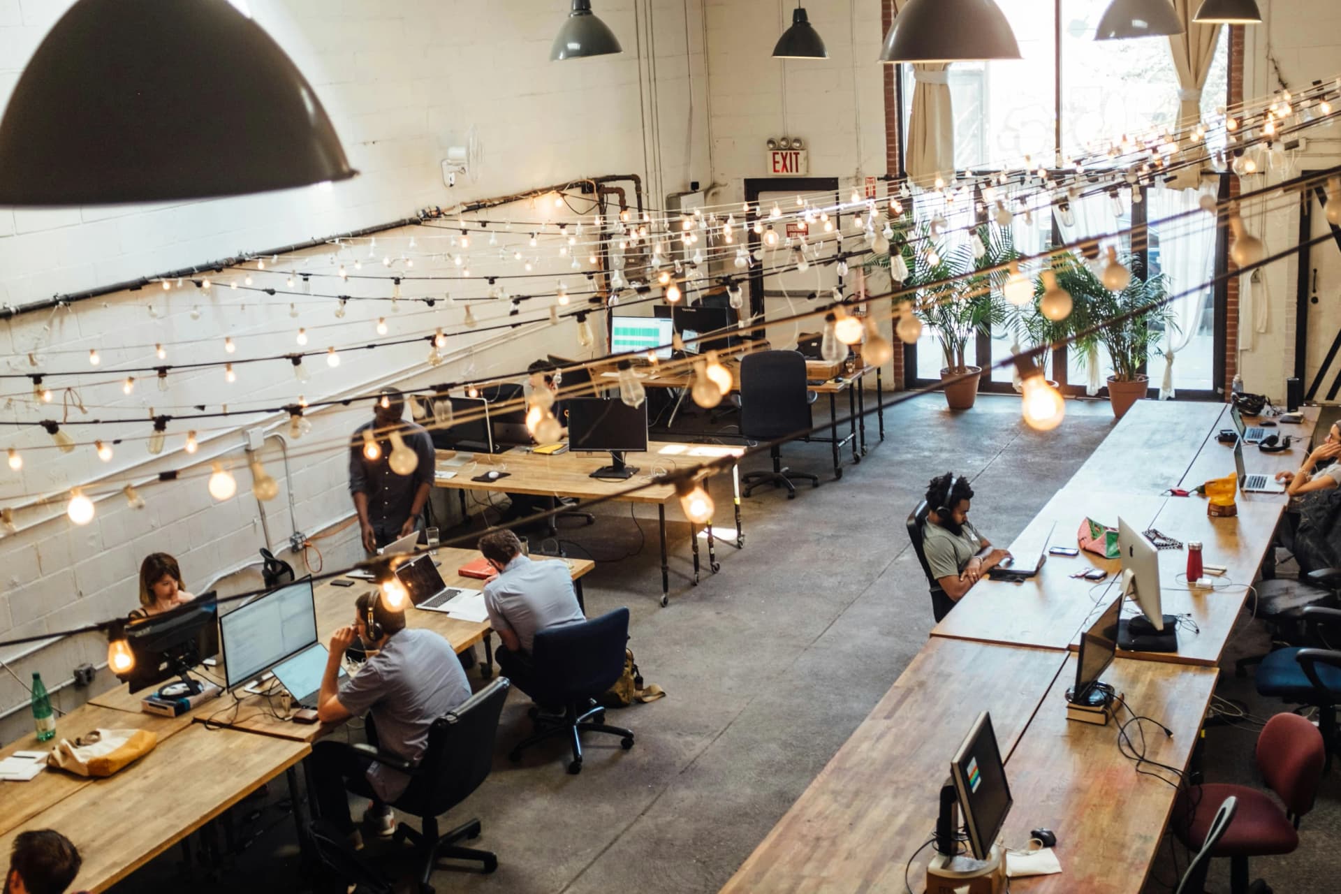 Overhead view of a busy industrial-style shared office with long wooden desks, monitors, pendant lamps, and strings of lights