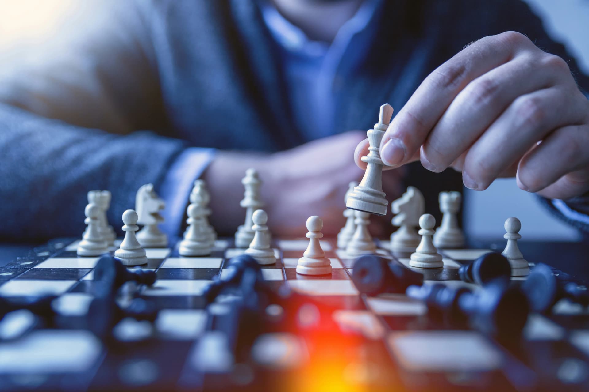 Hand moving a white chess piece across a board, with fallen dark pieces in the blurred foreground