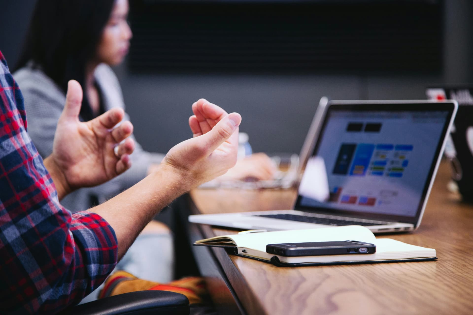 Person gesturing with both hands during a meeting at a table with a laptop and notebook, while a colleague works in the background