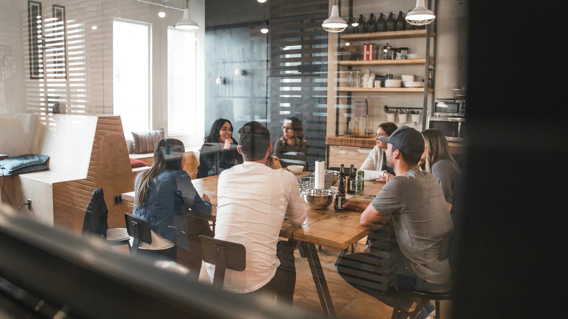Group of colleagues sitting around a large wooden table in a bright, loft-style office kitchen, seen through a window