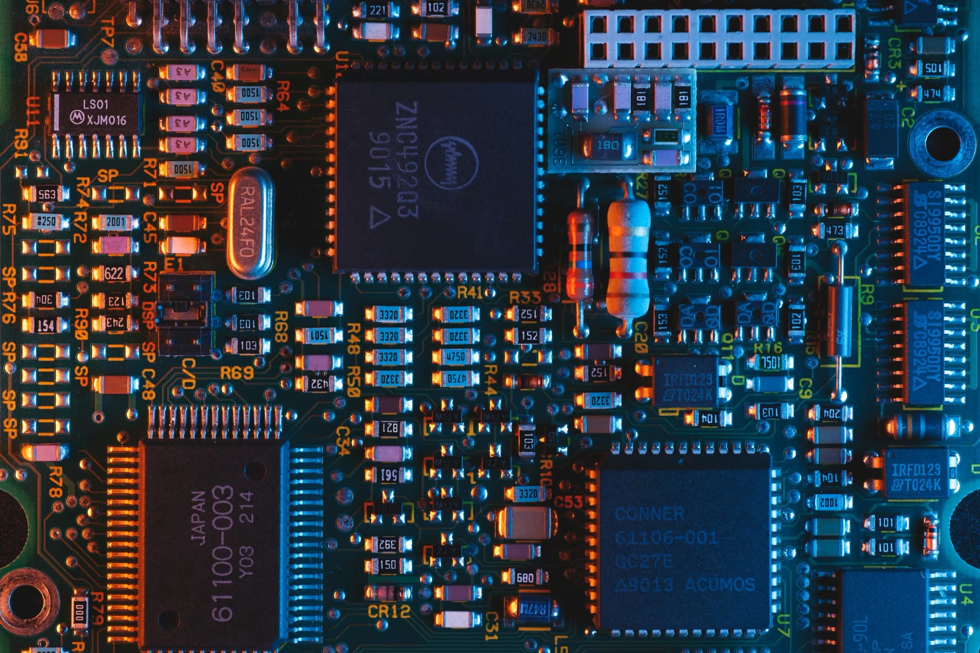 Close-up of a green printed circuit board with integrated circuit chips, resistors, and capacitors lit in blue and orange
