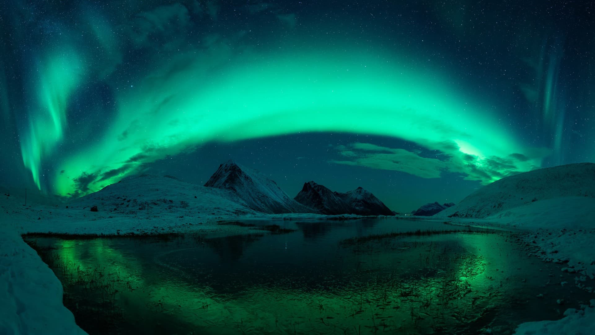 Green northern lights arching over snowy mountains and reflecting in a partly frozen lake under a starry night sky