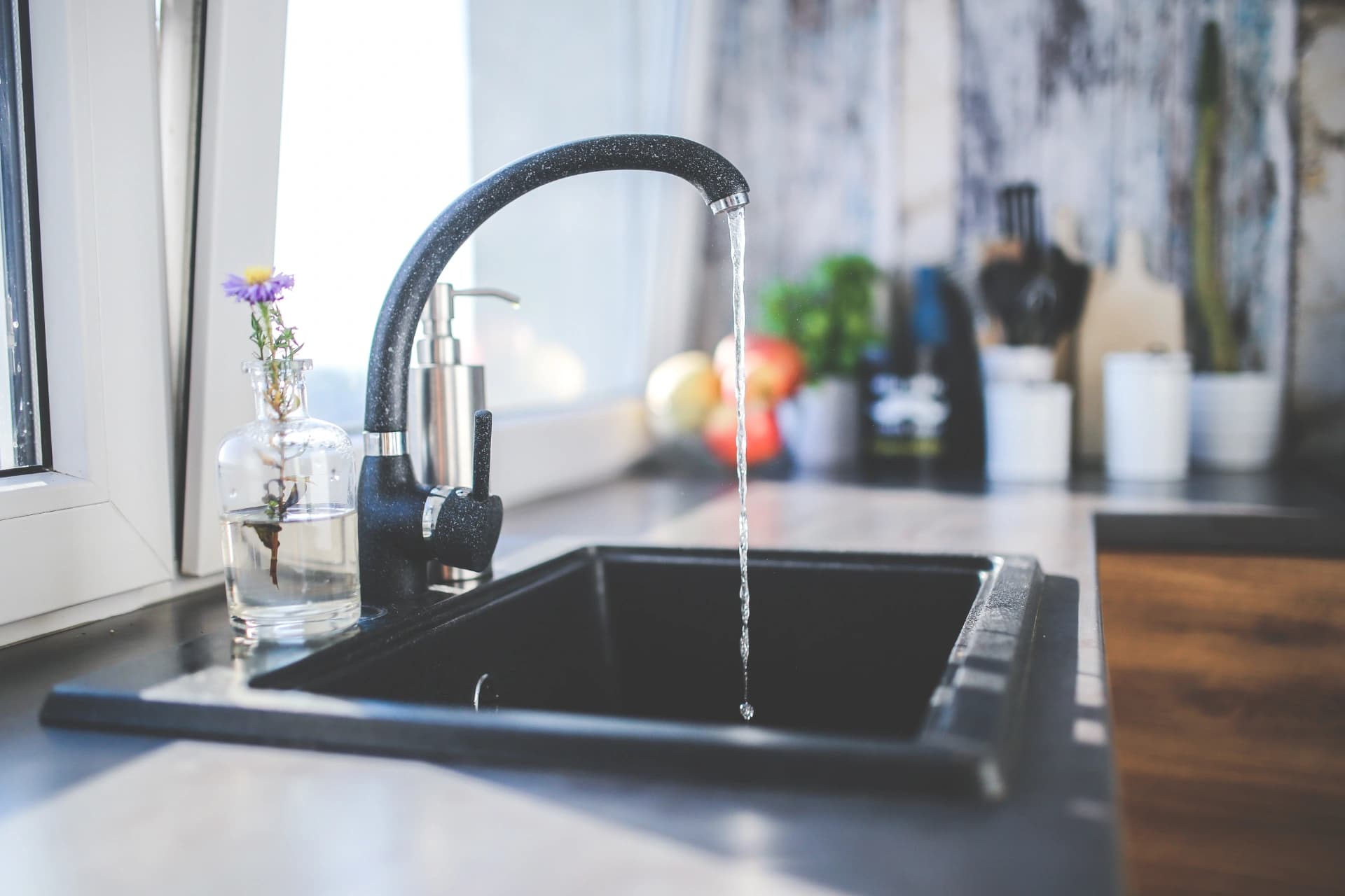 A black kitchen faucet running a thin stream of water into a black sink, beside a small glass vase holding a purple flower