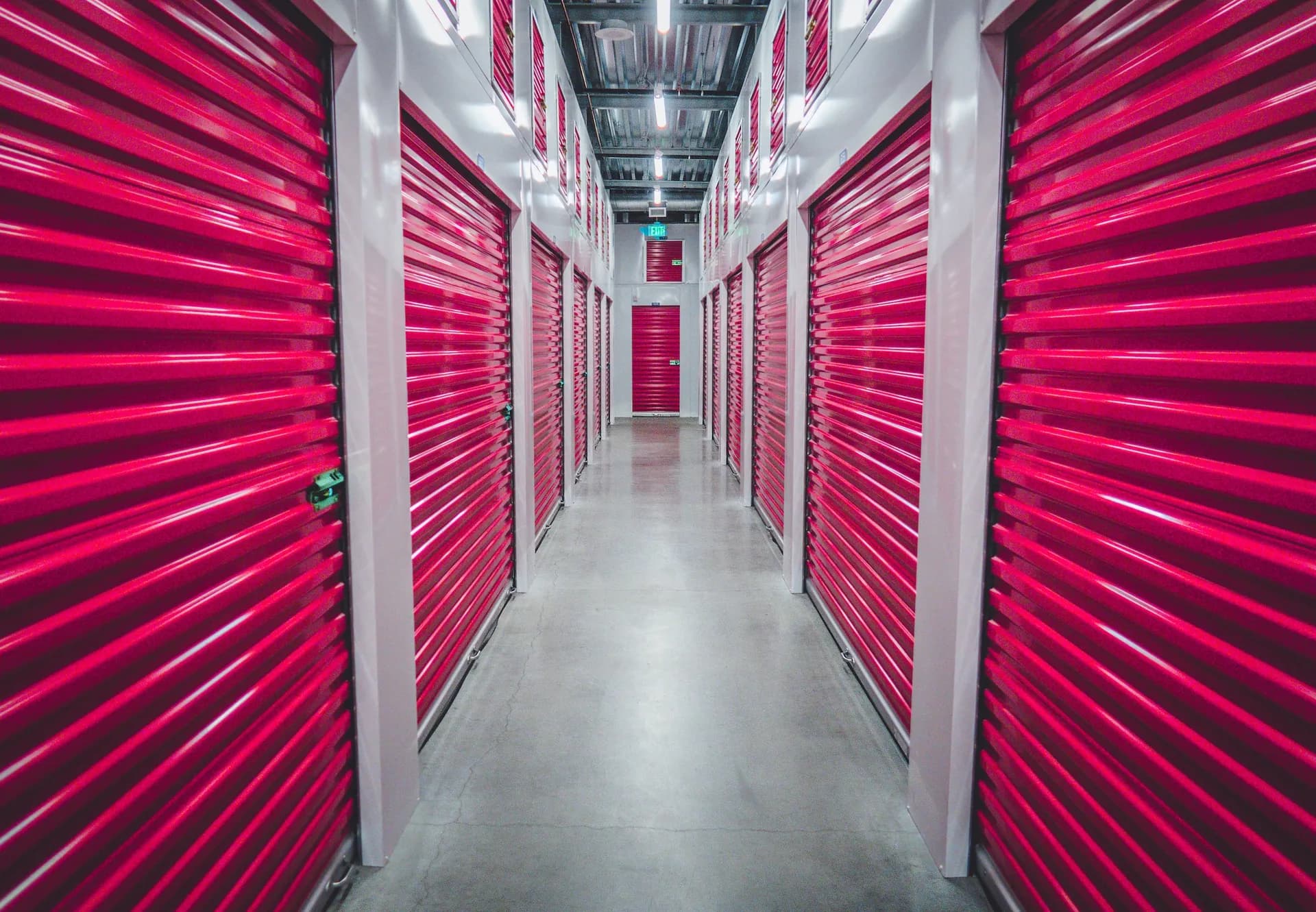 A long indoor corridor lined on both sides with bright red roll-up doors of self-storage units, ending at a lit exit sign
