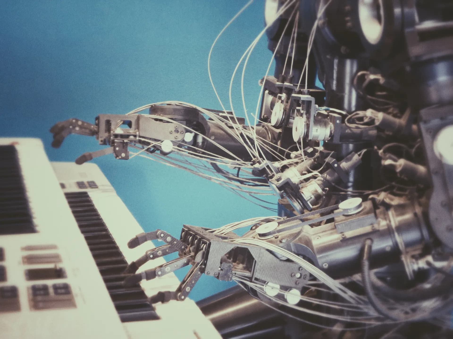 Robot with wire-strung mechanical hands playing an electronic keyboard against a blue background