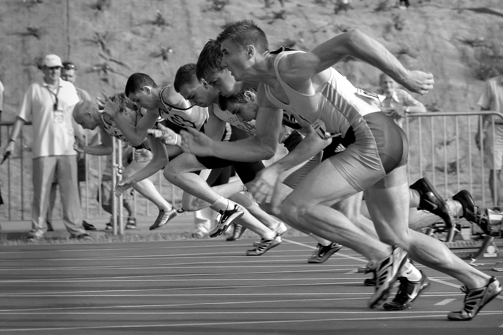 Black-and-white photo of sprinters exploding out of the starting blocks on a running track as officials watch from behind a fence