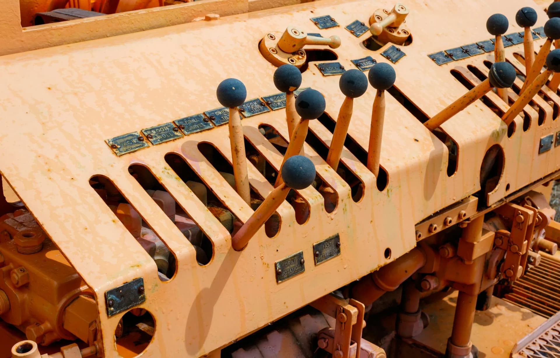 Close-up of a weathered orange machine control panel with rows of ball-topped levers and metal labels such as Drill Rotation, Boom Swing, and Fast Retraction