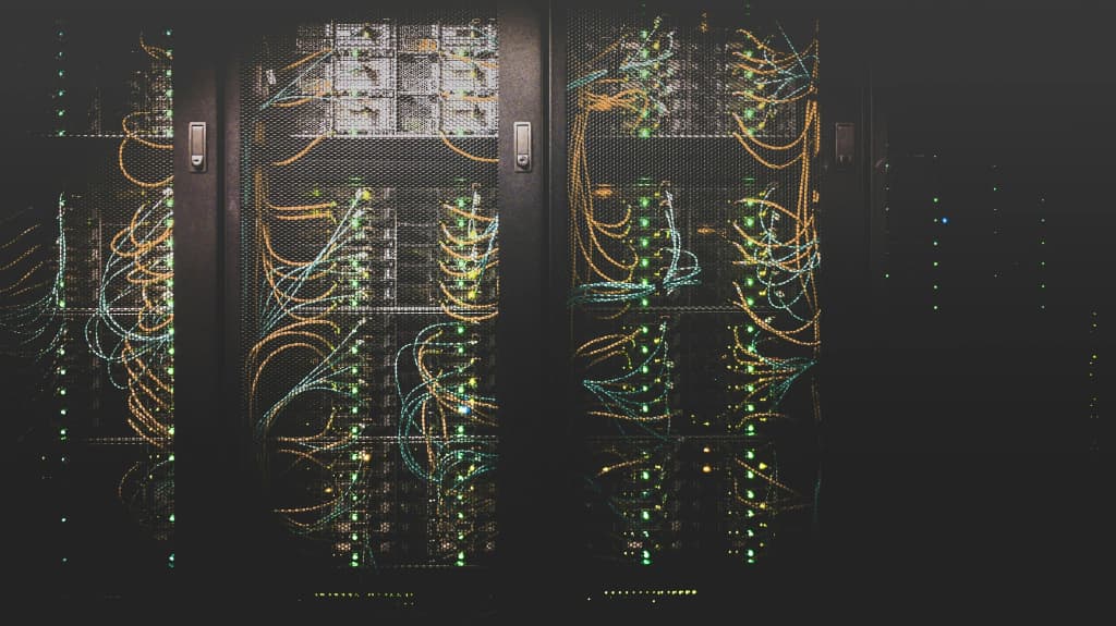 Dark server racks behind mesh doors, filled with tangles of orange and teal network cables and green status lights