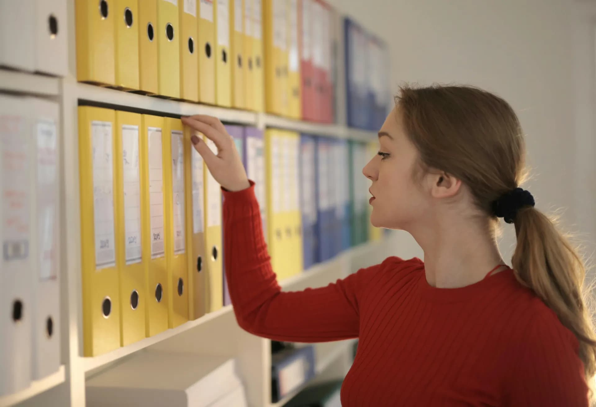 Woman in a red sweater pulling a yellow binder from a shelf of color-coded office binders