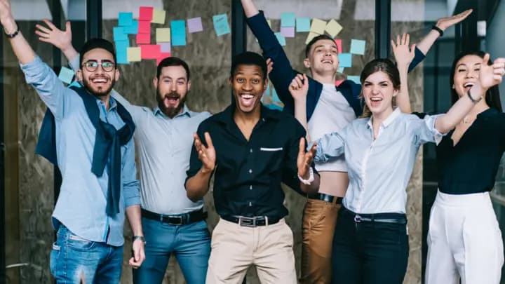A group of six smiling coworkers cheering with raised arms in front of a glass wall covered in colorful sticky notes