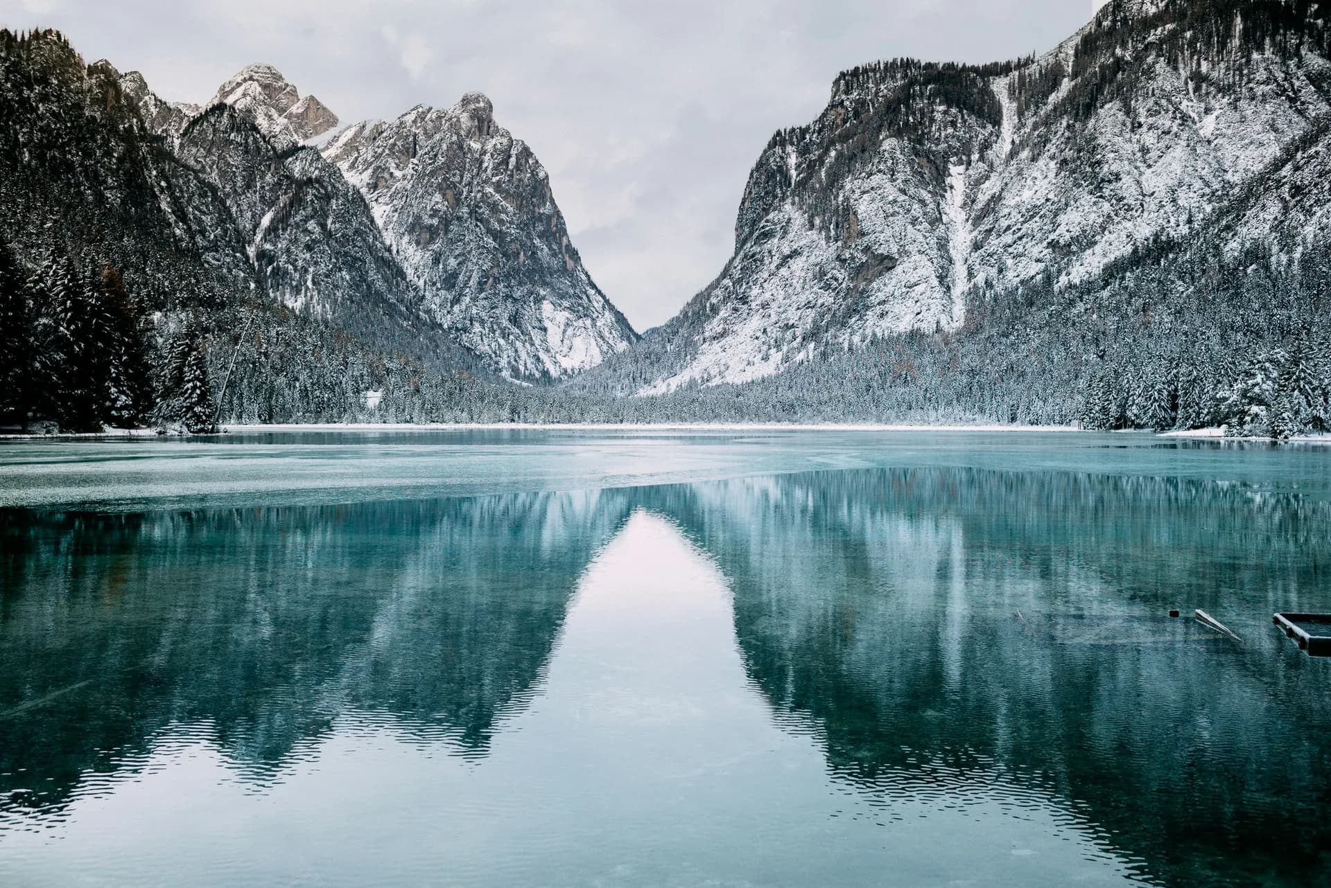 Snow-dusted mountains and evergreen forest reflected in the still turquoise water of an alpine lake under an overcast sky