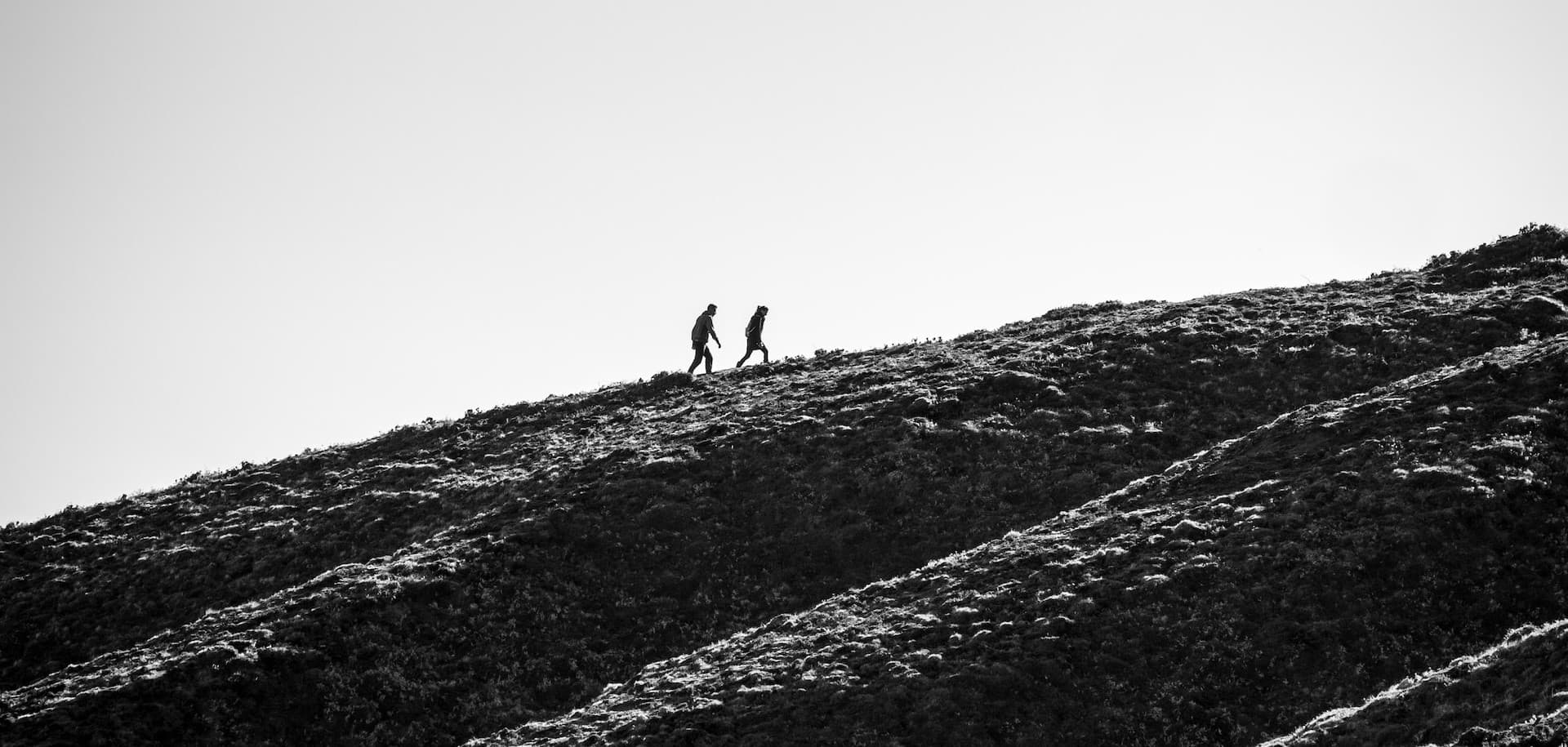 Black and white photo of two people hiking up a steep, grassy hillside, silhouetted against a pale sky
